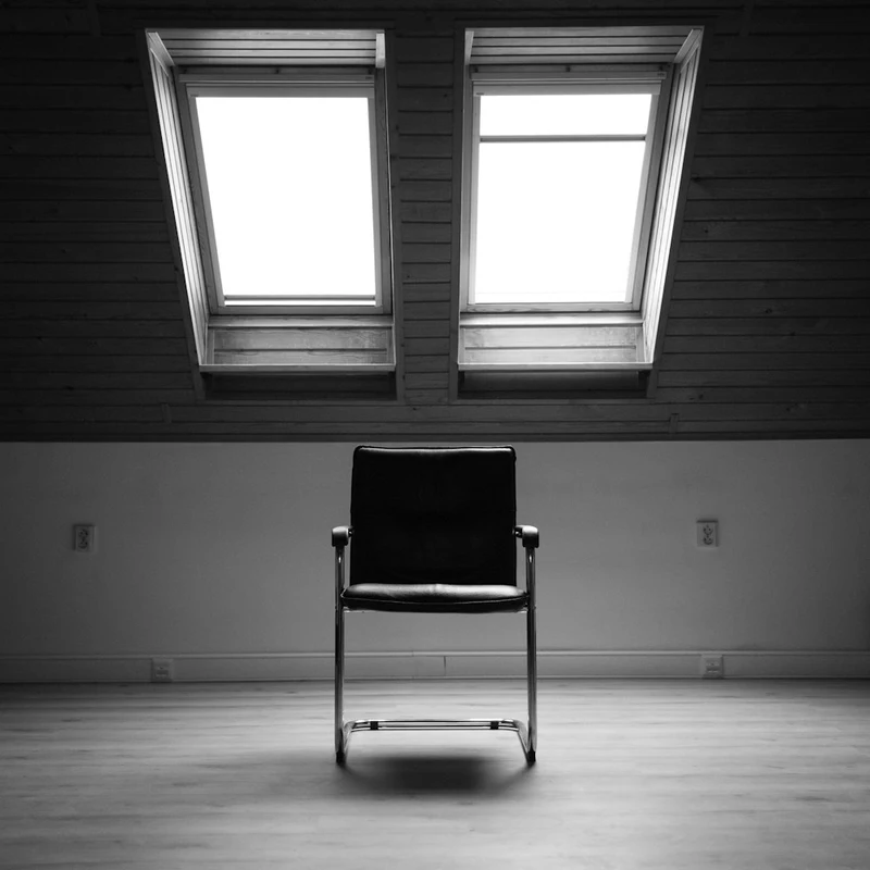 Chaise vide sous deux fenêtres de toit, pièce sobre en noir et blanc